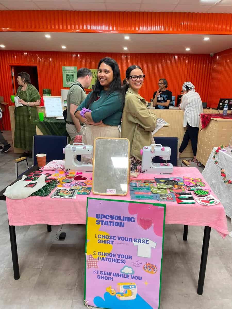 Two smiling women at an upcycling station with sewing machines and fabric patches, promoting sustainable fashion and animal rights awareness.